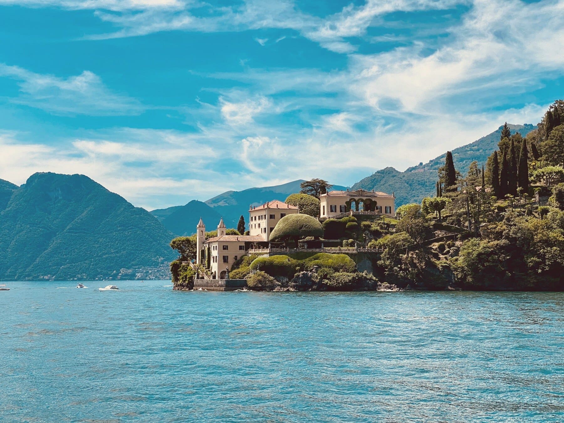 A villa and its gardens on a wooded promontory on Lake Como, mountains rising behind the far shore.