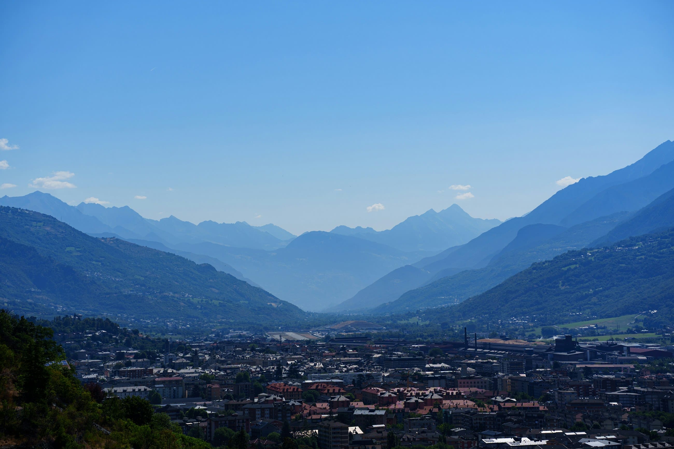 An Alpine valley in afternoon haze, the town spread along its floor and ridges receding behind.