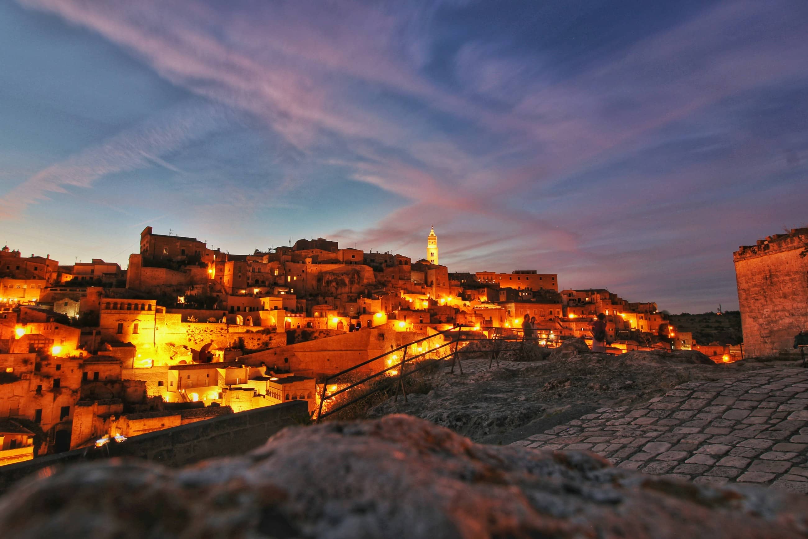 The sassi of Matera lit gold after sunset, stone houses stacked up the hillside under a violet sky.