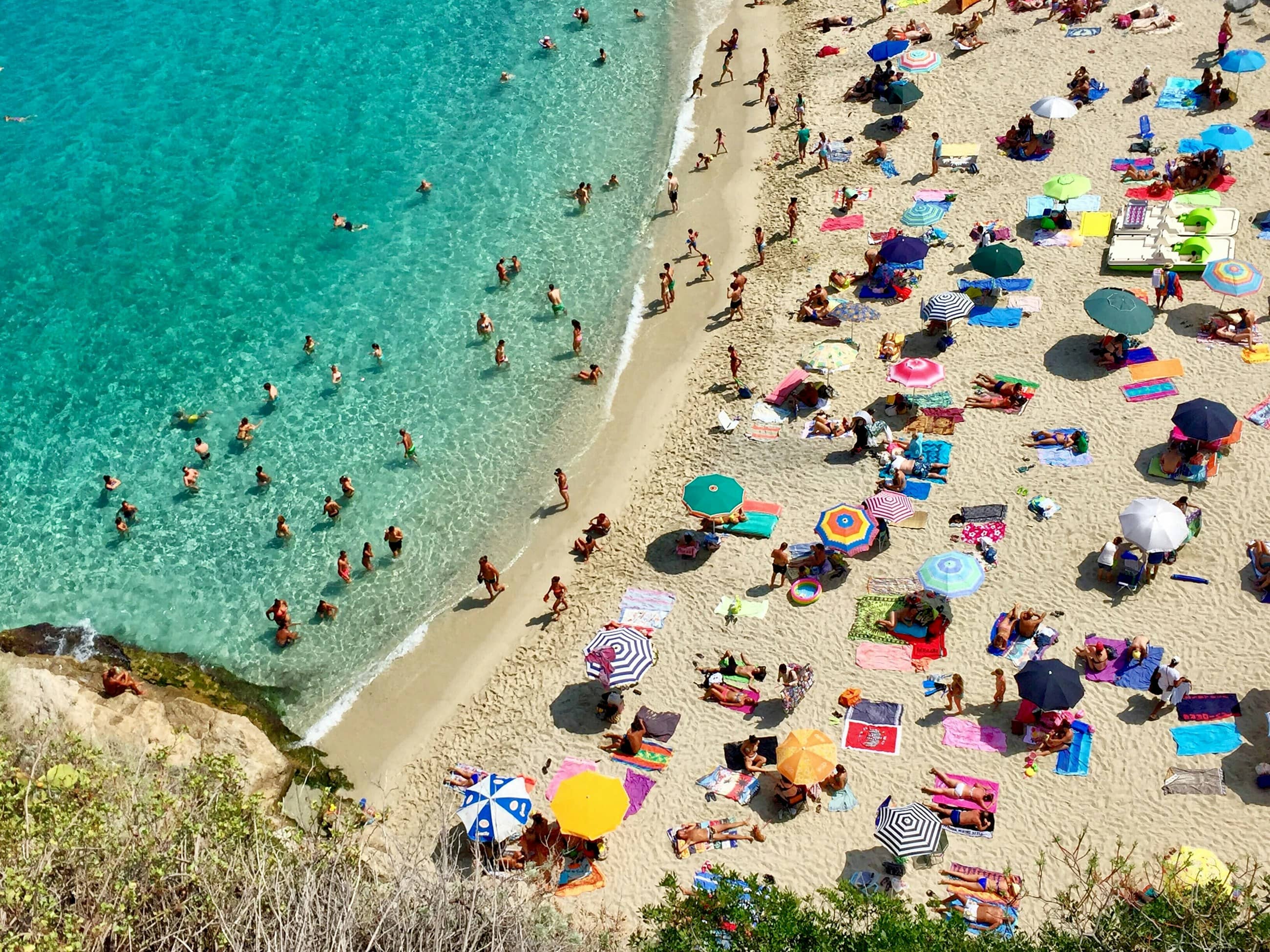 Looking straight down onto a Calabrian beach crowded with coloured parasols, turquoise water running along the sand.