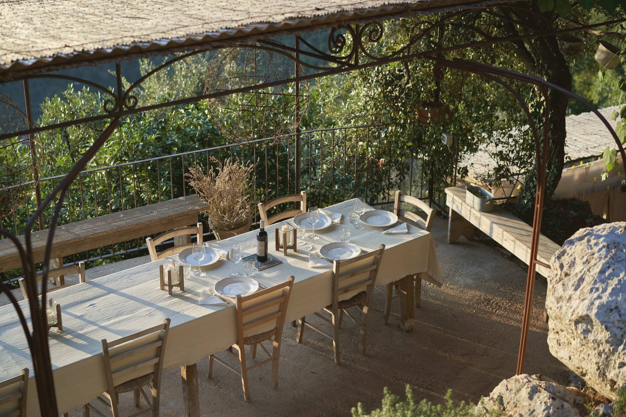 One long table laid under a vine pergola, benches down both sides, the garden bright behind it.