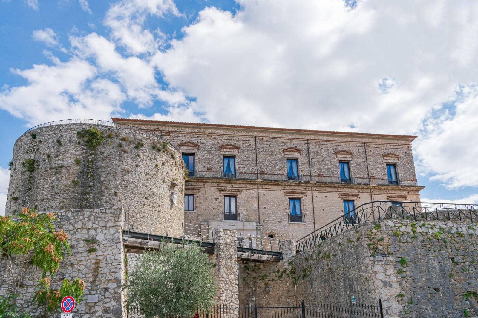 Castello Macchiaroli, Teggiano, Campania: exterior, the castle block and its round tower seen from below the ramparts against a bright cloud sky.