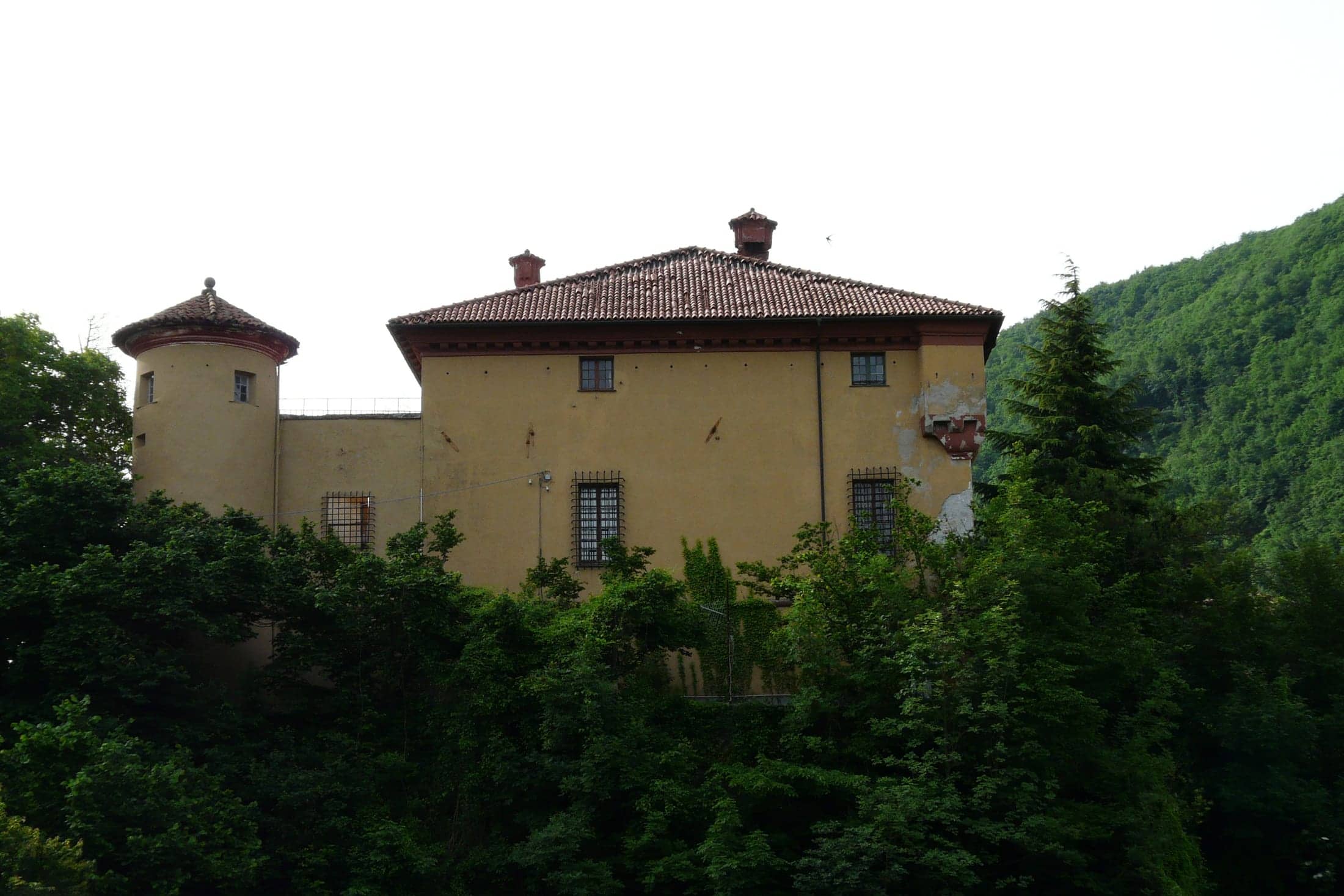 Castello Spinola Mignacco (trades as 'Castello Spinola'), Isola del Cantone, Liguria: exterior: the square manor block with its round corner tower on the wooded rock spur, seen across the treetops, landscape orientation.
