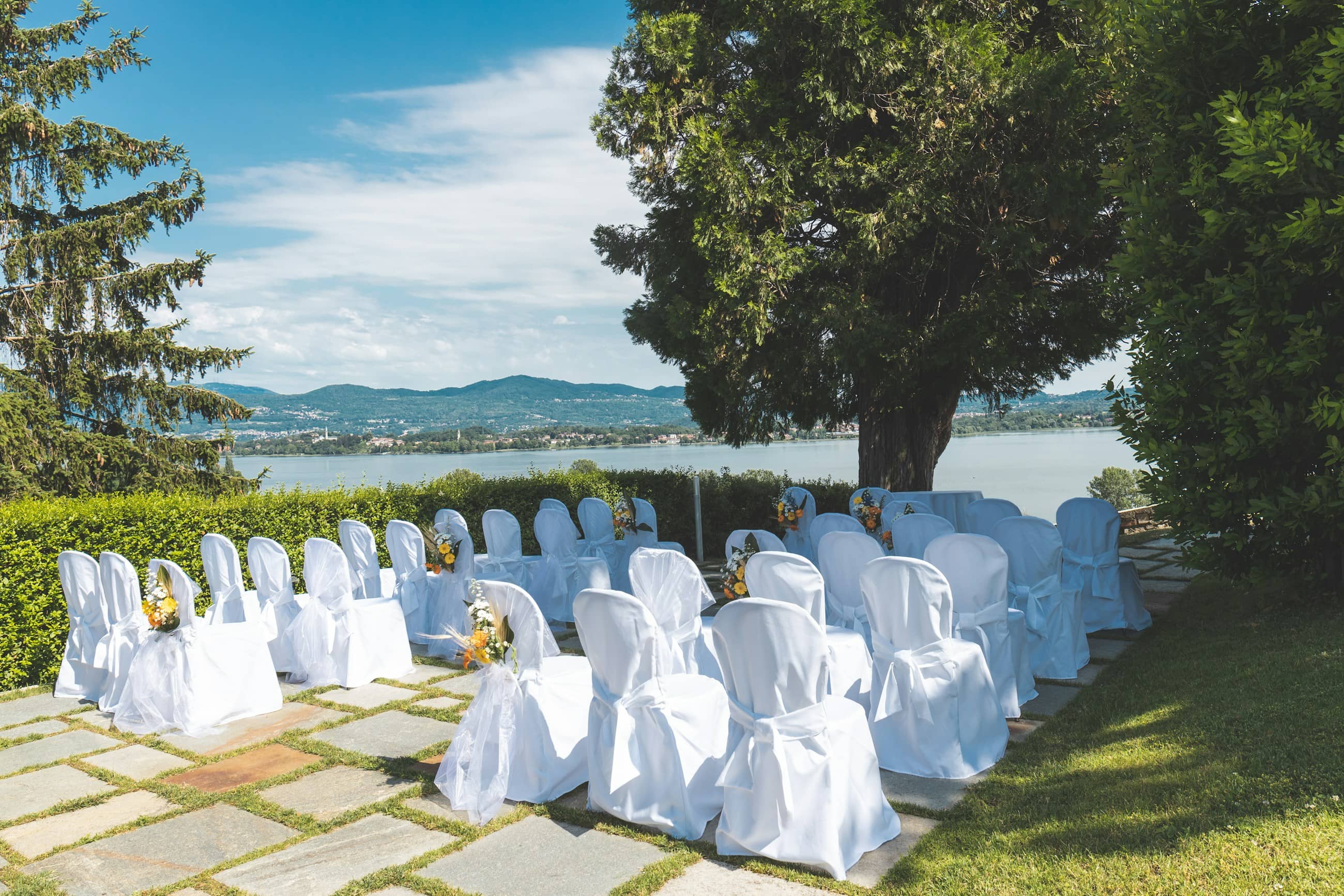 Rows of white ceremony chairs under trees beside the water, before the guests arrive.
