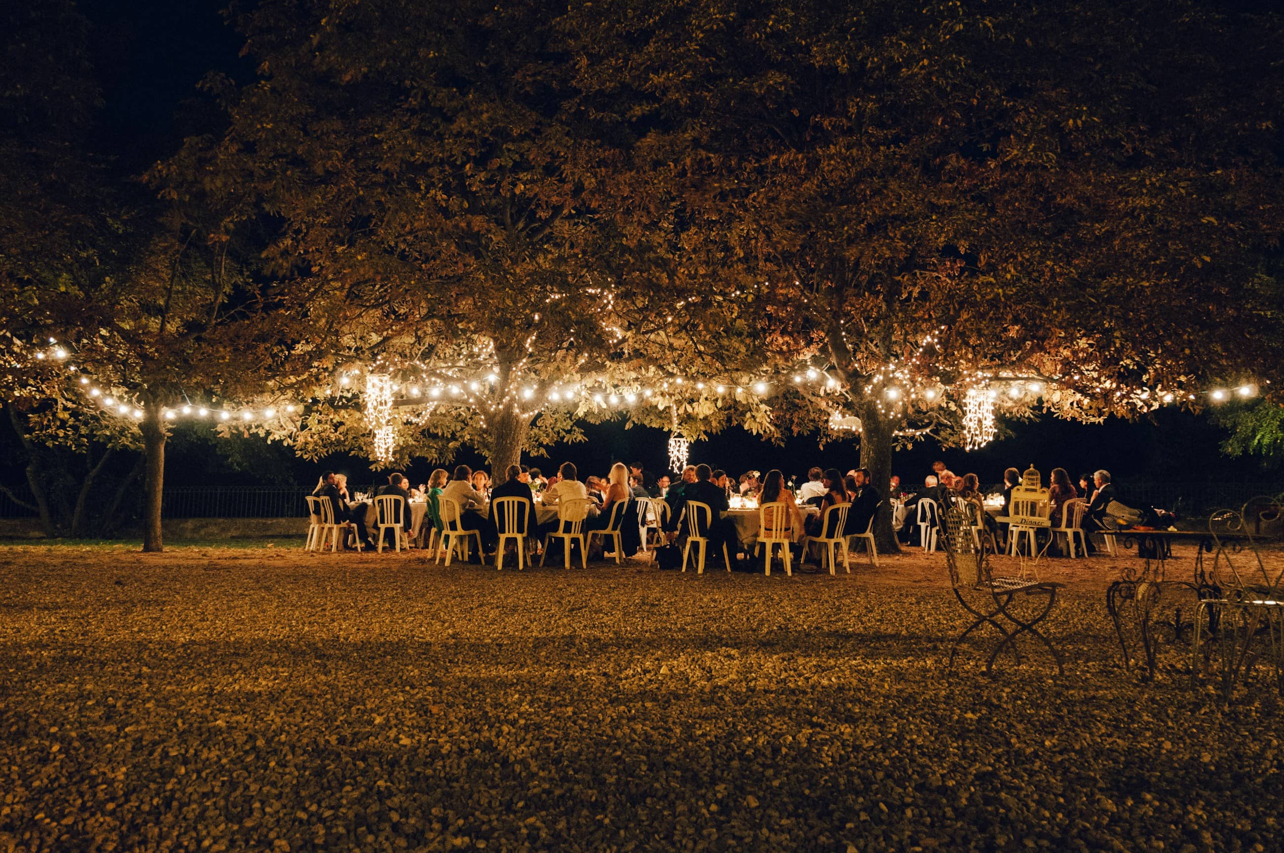 A wedding party at long tables under trees strung with lights, at night.